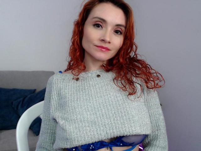 Drikka met rood krullend haar, die een grijze gebreide trui draagt, waarbij een blauwe bh en borsten zichtbaar zijn, zittend op een witte stoel, met een lichte glimlach, roze lippenstift, bleke huid, informele binnenomgeving..