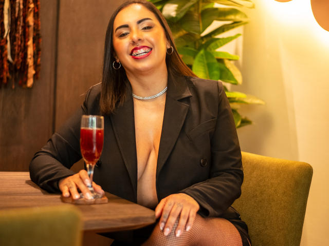 Genexis with long black hair, smiling, wearing a black blazer, exposing her big tits, holding a red wine glass, sitting at a wooden table, wearing fishnet stockings.