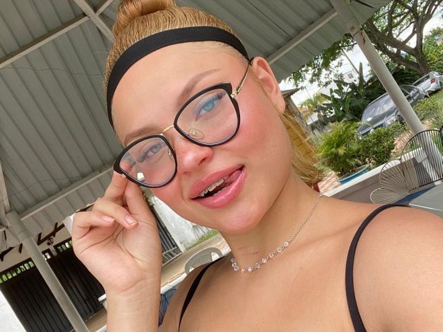 Meean with blonde hair in a bun, black headband, and glasses, tongue slightly out, wearing a black strap top and a delicate necklace, smiling at the camera.