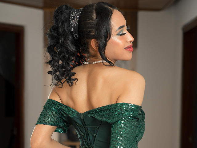 MichellDiaz a les cheveux noirs bouclés, porte une robe verte pailletée à une épaule, un collier de perles et du maquillage blanc sur les joues. Elle est de profil, montrant son dos nu et ses lèvres rouges..