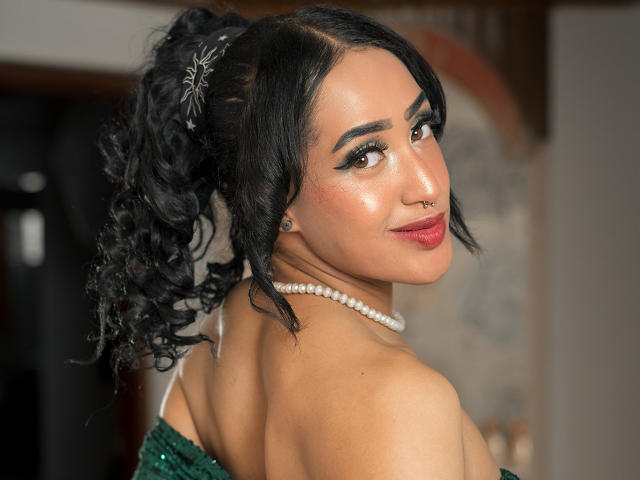 MichellDiaz, con i capelli neri ricci raccolti in una coda di cavallo, indossa un abito verde con una spalla scoperta, una collana di perle, un anello al naso e un trucco occhi smokey, sorridendo di spalle. Il seno è parzialmente visibile..
