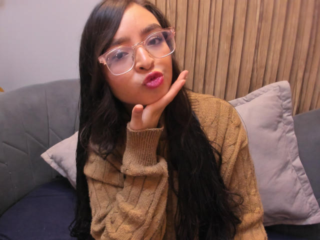 EvaMcAllister with long black hair, pink lips, clear glasses, and a brown knitted sweater, pouting with her hand on her cheek, sitting on a gray couch.