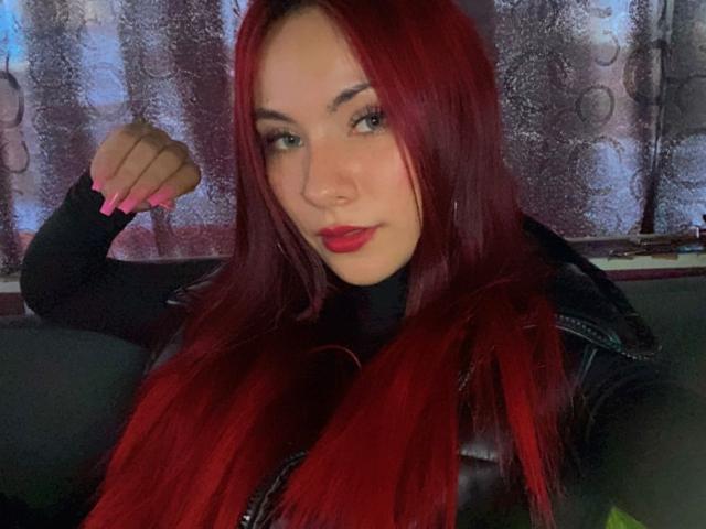 ValeryHarllow with red hair, black turtleneck, and black leather jacket, has pink nails and red lipstick, posing with a slight smile, right arm raised.