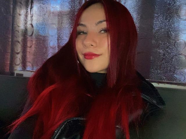 ValeryHarllow with long red hair, wearing a black jacket, has red lipstick and a slight smile. Her tits are partially visible. She's sitting in a dimly lit room.