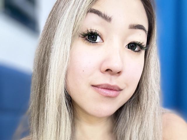RenyLime hat blondes Haar, schwarzen Eyeliner, lange Wimpern und rosafarbene Lippen. Sie schaut mit einem leichten Lächeln in die Kamera. Ihre Haut ist hell. Sie hat kleine Sommersprossen..