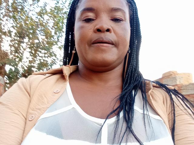 MelonBoobsI is a chubby Black woman with long braids, wearing a beige shirt over a white mesh top, showing her small tits, with a neutral expression, outdoors.