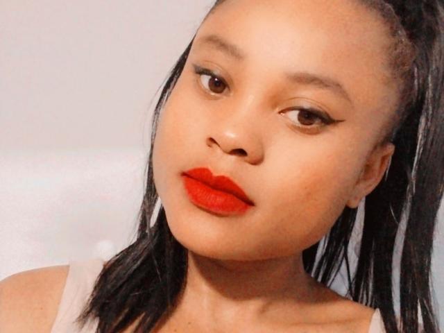 NehalScott has smooth brown skin, red lipstick, black eyeliner, and black high-ponytail. She's looking to the side with a slight pout, wearing a beige top.