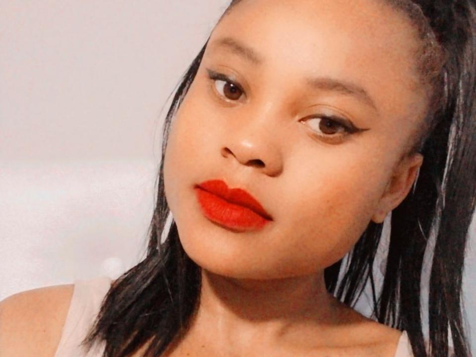 NehalScott has smooth brown skin, red lipstick, black eyeliner, and black high-ponytail. She's looking to the side with a slight pout, wearing a beige top.