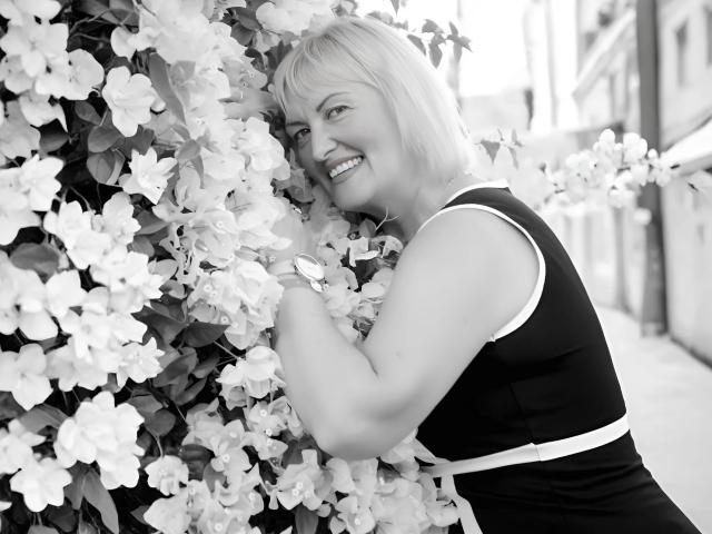 SecretFoxy with blonde hair, smiling, hugging a wall of blooming flowers, wearing a sleeveless black dress with white trim, standing outside on a sunny day.