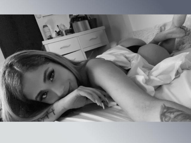 Black-and-white photo of KattyWattson lying on bed, looking at camera with a smile. Her ass is visible, covered by a white sheet. She has a cross tattoo on her wrist.