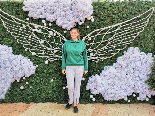LustCaroline stands in front of a green wall with white rope wings and purple clouds, wearing a green shirt and light purple pants, smiling with short brown hair.