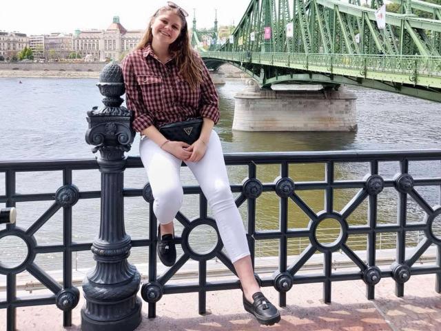 LustCaroline, with long brown hair, sits on a black iron railing, wearing a red check shirt, white pants, black shoes, and a black belt, smiling at a green bridge.