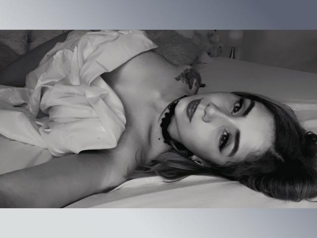 KattyWattson is lying on a bed, topless with a white shirt partially covering her tits, wearing a choker, looking at the camera with dark eyebrows and a slight smile.