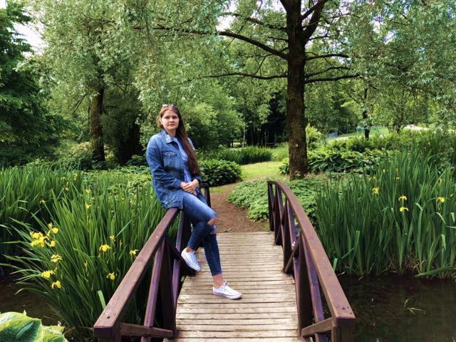 LustCaroline sitting on a wooden bridge, wearing a denim jacket and blue jeans, with white sneakers, and a slight smile, surrounded by greenery and yellow flowers.