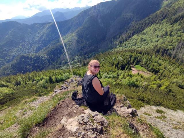 LustCaroline with her ass facing the camera, sitting on a rocky mountain trail in black clothes and sunglasses, looking back at the camera. Forest and mountains in background.
