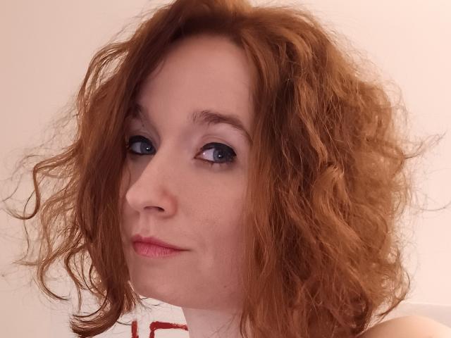 SexyGoth69 has curly red hair, blue eyes, and pink lips. She's looking to the side with a slight smile, showing a cute, confident face. Her tits are small.