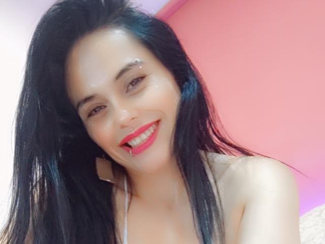 CharlotteCrofort with shiny face, black hair, and red lipstick smiling at the camera, showing her teeth and small nose, wearing a halter top, in a pink-lit room.