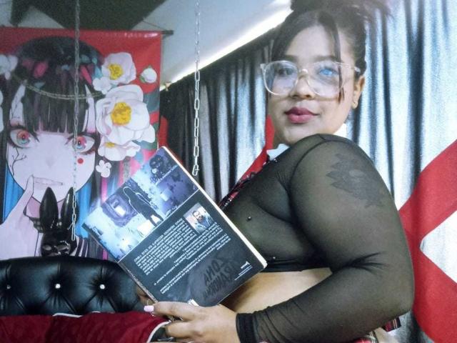 PamelaMckein with glasses, black sheer top, holding book, showing her tits, dark hair in bun, smiling, black cat hanging, red and white flag in background, tufted black chair.