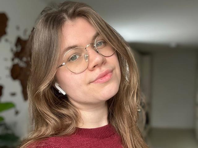 LinaLove: una chica atractiva con cabello castaño ondulado, gafas y un suéter rojo, sonriendo a la cámara, con auriculares blancos, luciendo adorable..