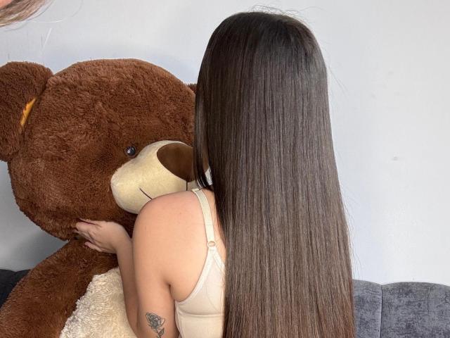 KatyJonnes with long brown hair, hugging a brown teddy bear, wearing a beige bra, visible tattoo on left arm, standing in front of a white wall.