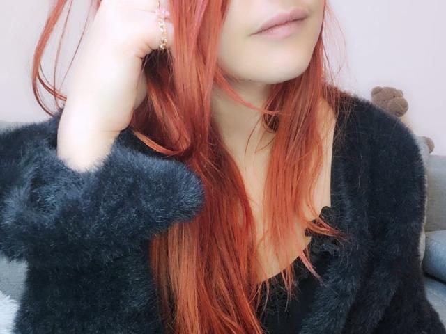 LilouLove with red hair, wearing a black fuzzy jacket, has her hand in her hair, showing a subtle smile, and a small ring on her finger.
