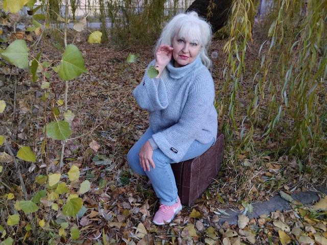 KamaliaSim with white hair, gray turtleneck, blue jeans, and pink sneakers, squatting on a brown box, touching her cheek, surrounded by yellow leaves and green vines.
