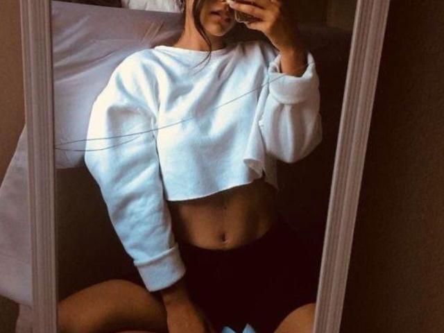 Mirror selfie of SweetAmbers with a white, long-sleeve crop top, exposing a flat, toned midriff, and black shorts, holding a phone, with her ass partially visible.
