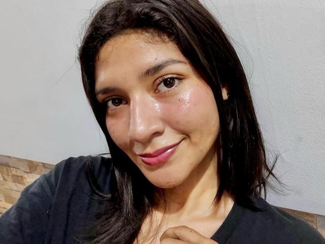SweetAmbers has shiny, sweaty skin, dark brown eyes, and a small smile. She's wearing a black shirt with her hand on her chest. Her pink lips are slightly parted.