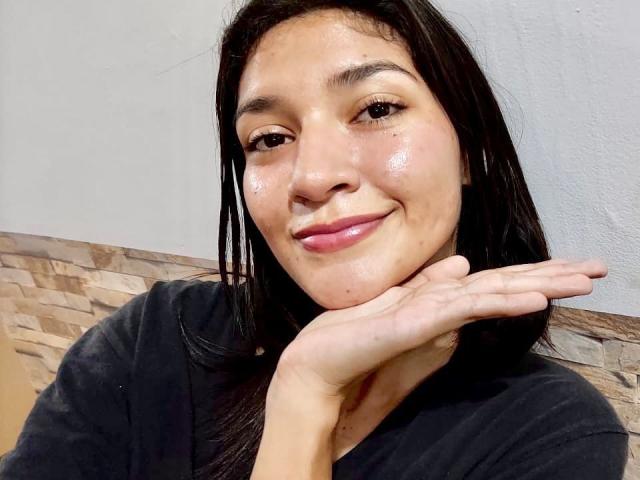 SweetAmbers has shiny skin, black hair, and a slight smile, resting her chin on her hand. She's wearing a black shirt, and her pink lips are visible.