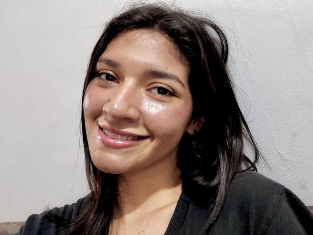 SweetAmbers has shiny, wet-looking skin, a cute smile with slightly parted pink lips, and long, messy black hair. She's wearing a black shirt, and her ass is not visible.