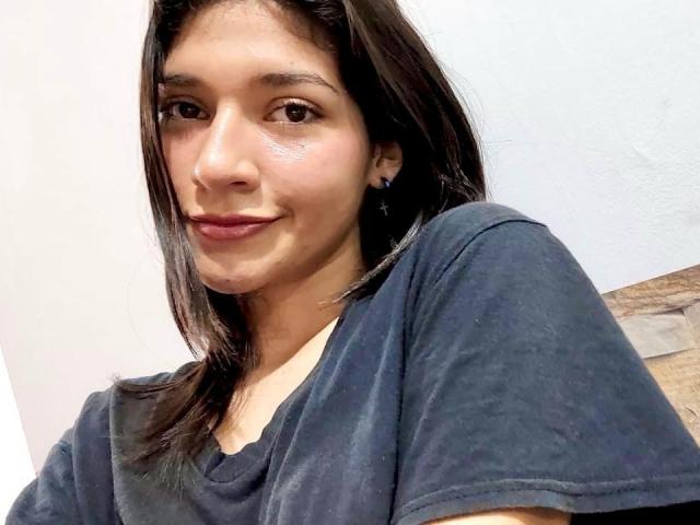 SweetAmbers with medium brown skin, black straight hair, and small tits, wearing a dark blue shirt, smiling slightly with pink lipstick, brown eyes, and subtle cheek glow.