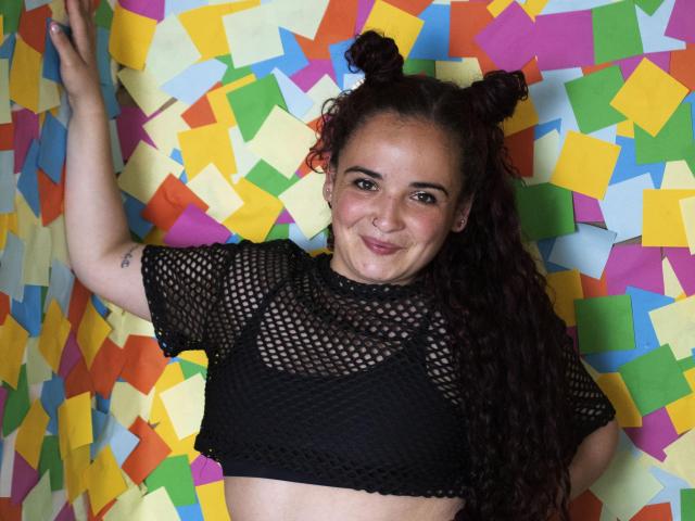 LunaF with curly dark red hair in double buns, smiling, wearing a black mesh crop top, exposing her small tits, against a colorful, patterned wall.