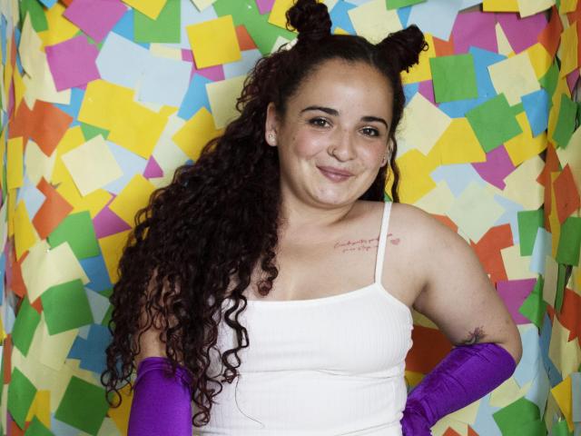 LunaF with curly brown hair in space buns, white tank top, purple gloves, small tattoo on right shoulder, smiling at camera, colorful paper background. Boobs visible, cute.