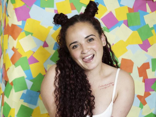 LunaF with curly brown hair in double buns, smiling, showing teeth, wearing a white tank top, small tits, tattoo on right chest, colorful background, cheerful expression.