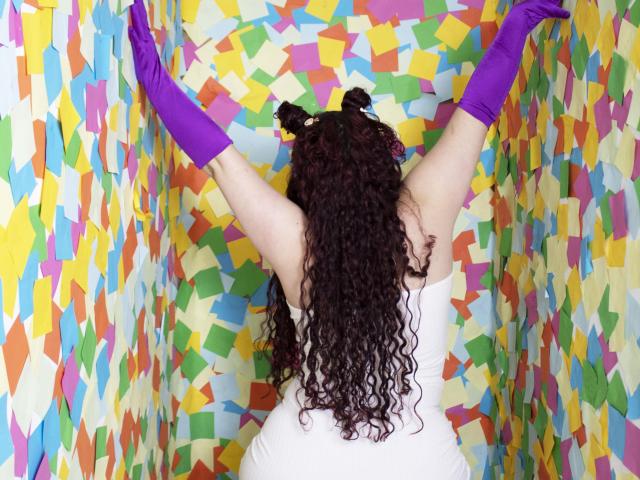 LunaF with curly dark hair, wearing a white dress and purple gloves, arms raised, back to camera, colorful paper strips in background. Boobs visible, ass not.