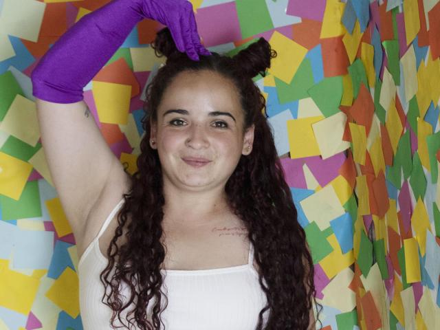LunaF with curly dark hair, white tank top, purple glove, holding double bun hairstyle, smiling, small tattoo on right chest, light skin, medium breasts, colorful paper backdrop.