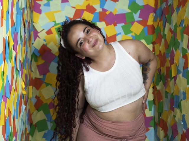 LunaF with curly brown hair, white ribbed tank top, pink skirt, smiling, hands on hips, visible tattoo on right arm, standing in colorful, abstract-patterned room.
