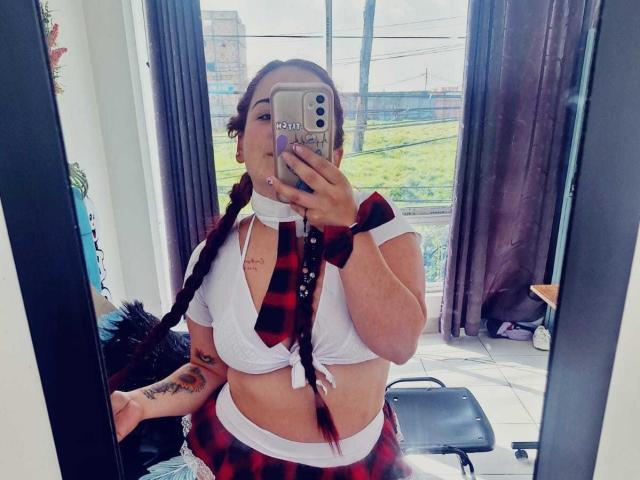 LunaF with braided red hair, white tied-up crop top, red plaid skirt, black wrist cuff, holding phone, mirror selfie, showing tits and a tattoo on her left arm.