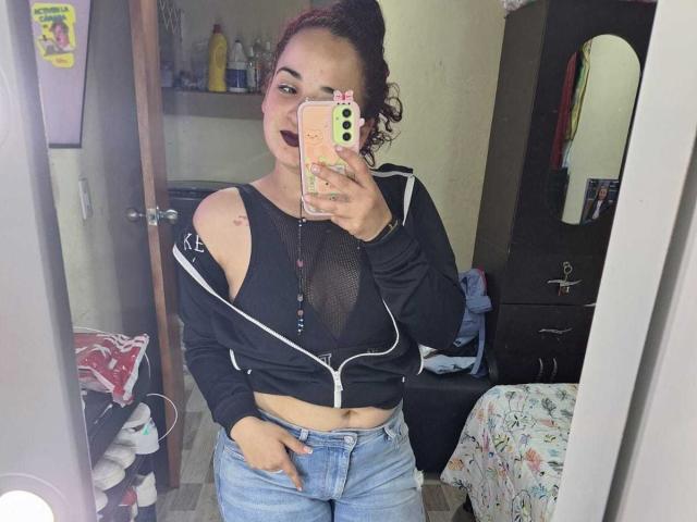 LunaF with dark makeup, wearing a black top with cutouts, showing her tits, and blue jeans, taking a mirror selfie in a bedroom, holding a pink phone.