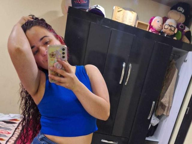 LunaF with red braided hair, blue crop top, and jeans, holding phone, showing armpit, and slightly covering tits, in a bedroom with black wardrobe and stuffed toys.