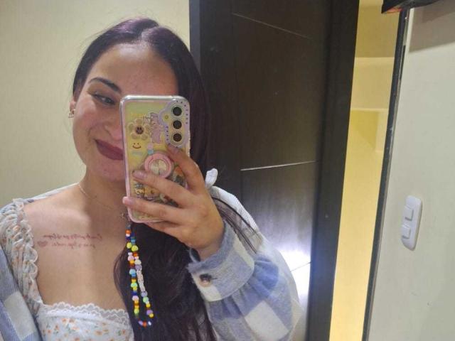 LunaF with long dark hair, smiling, holding phone with colorful case, wearing checkered shirt, lace dress, showing small tits, tattoo on right chest, in mirror selfie.