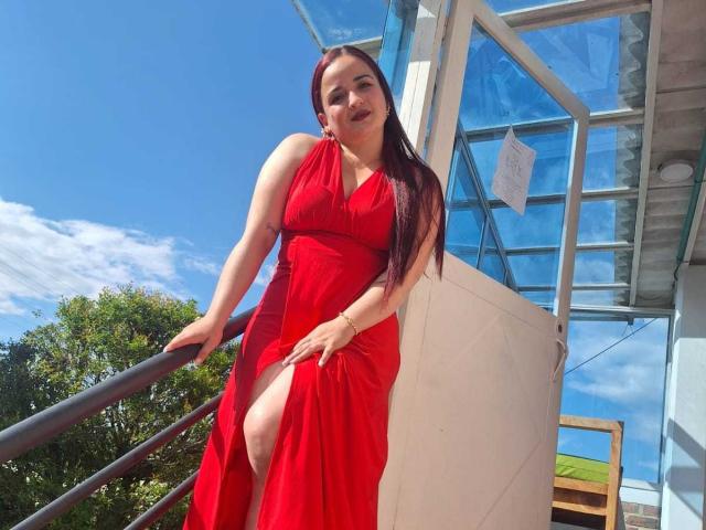 LunaF with long red hair, wearing a red dress with a thigh-high slit, standing on a metal staircase, blue sky, and glass building in the background.