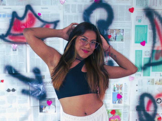 KimmySolange with glasses, black sports bra, white pants, hands in long brown hair, smiling, standing, graffiti in background, pink hearts, black spray paint, confident, cute, tattoo on right arm.