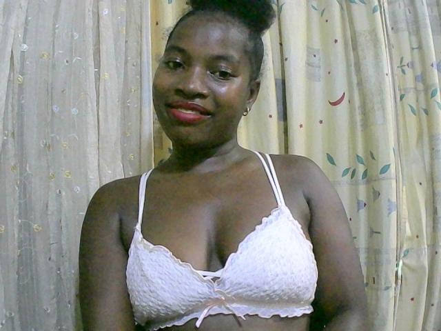  AlexandraC with dark skin, red lipstick, and a bun, wearing a white lace bra, smiling at the camera, showing her small tits. Curtains with a floral pattern in the background.