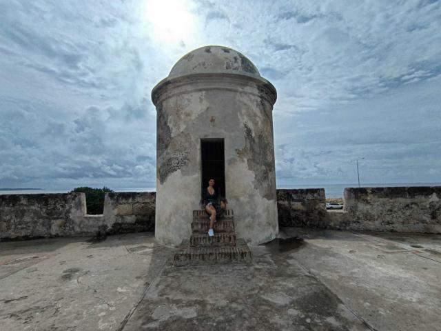 KatherinLatin, standing in front of a round, white, worn-down building with a dome roof, sits on stone steps, wearing a black shirt and shorts, under a cloudy sky.