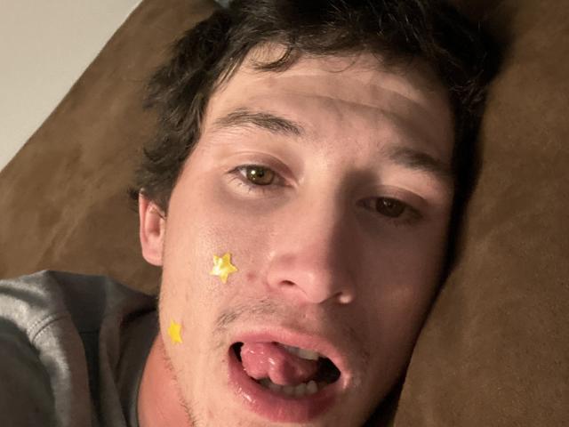 Le visage d'AlecurClit, des yeux marron, des cheveux foncés, une étoile jaune collante et un petit smiley jaune sur la joue gauche, la langue tirée, des lèvres roses, une bouche légèrement ouverte, allongé sur une surface marron..