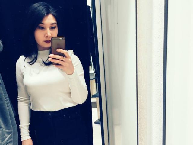 SoftGeraldine, avec ses cheveux foncés, portant un pull col roulé blanc et un pantalon noir, tenant un téléphone, prenant un selfie devant un miroir, montrant ses seins et ses fesses dans le reflet..