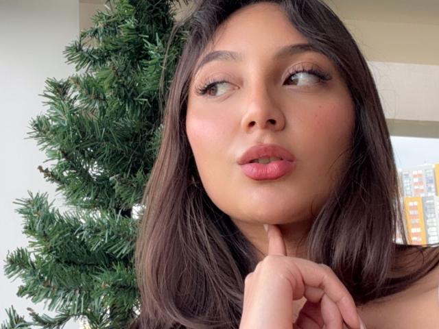 DianaFernandezz with brown hair, pink lips, and her finger near her chin, looking to the side, next to a green Christmas tree. Her face is slightly flushed.