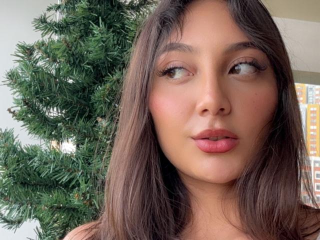 DianaFernandezz ha la pelle marrone media, i capelli lunghi castano scuro e le labbra rosate. Guarda di lato con gli occhi verdi. Sullo sfondo si vede un albero di Natale verde..