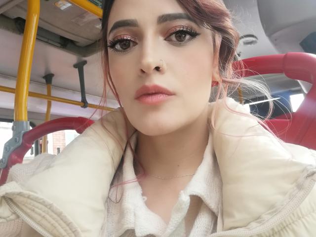JaneRoa with pink eyelids, long eyelashes, and pink lipstick, wearing a beige jacket over a white shirt, inside a yellow and red bus. She has a nose ring.
